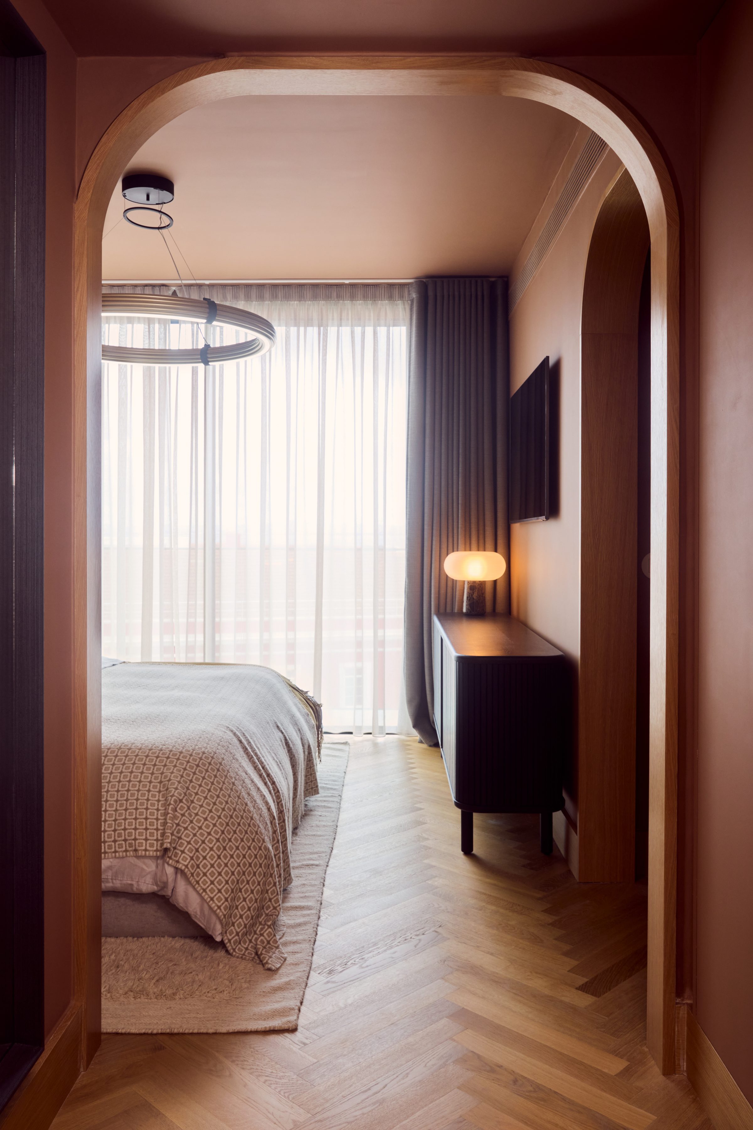 A room by Matt Smith-Wood in shadow, seen through an arched oak doorway, with daylight coming through a sheer-curtained window