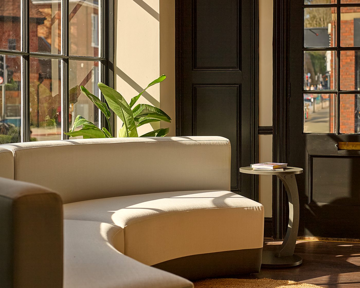 The waiting room at Sona Dental: late sun across a curved cream sofa, a jute rug and the street beyond tall Crittall glazing