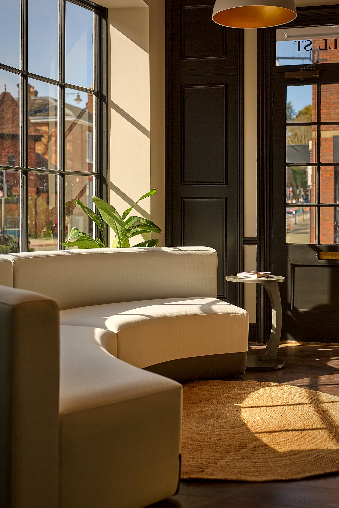 Late afternoon sun falling across a curved cream sofa and a jute rug in the waiting room at Sona Dental, with the street visible through tall Crittall glazing